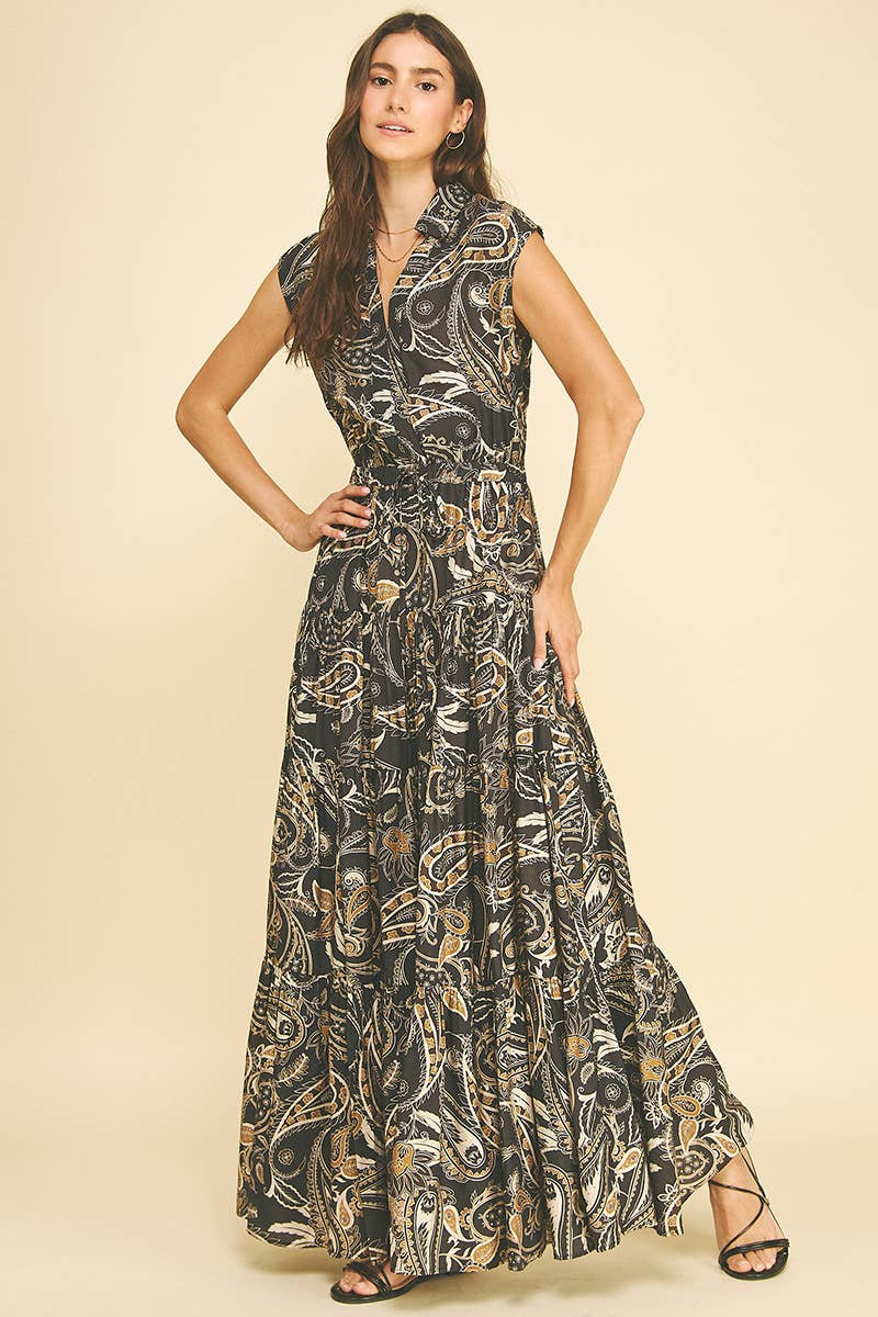 A woman modeling a sleeveless, floor-length wrap dress with a black base and an intricate beige and gold abstract paisley pattern, set against a solid light beige background.