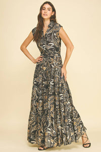 A woman modeling a sleeveless, floor-length wrap dress with a black base and an intricate beige and gold abstract paisley pattern, set against a solid light beige background.