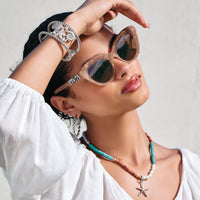 Model wearing Brighton cat-eye sunglasses with blush frames, styled with a silver starfish cuff and beaded necklace